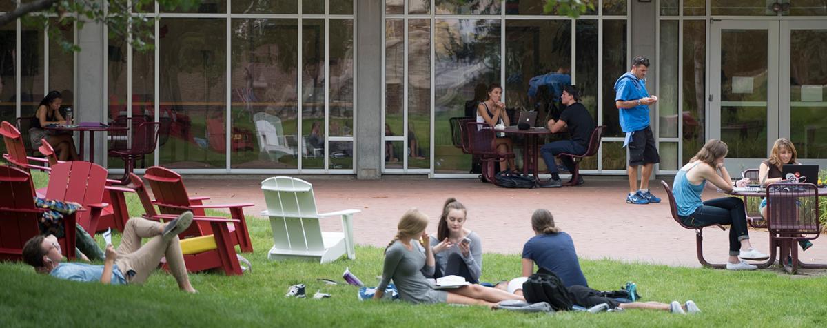 group of students lounging on grass 和 sitting at tables outside glass-walled building